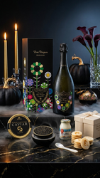 A luxury gourmet gift set arranged on a dark marble surface against a dark background. The set includes a bottle of Dom Pérignon Champagne, its floral illustrated box, a tin of caviar, a small jar, cookies, a spoon, and a gift box, surrounded by decorative props like lit candles, black pumpkins, and calla lilies.