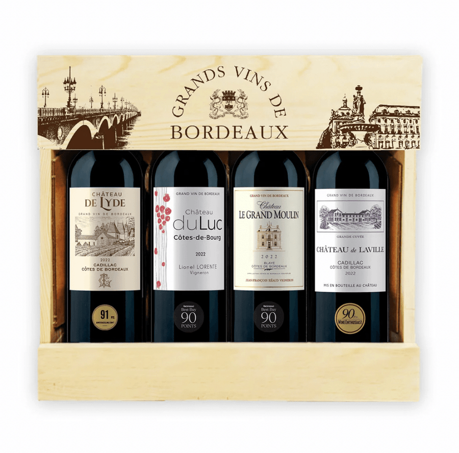 Wooden gift crate containing four 75cl Bordeaux red wine bottles neatly arranged in a row, presented as a premium Bordeaux wine gift pack. The light natural wood box has a hinged lid (open) with burnt-in branding: "GRANDS VINS DE BORDEAUX