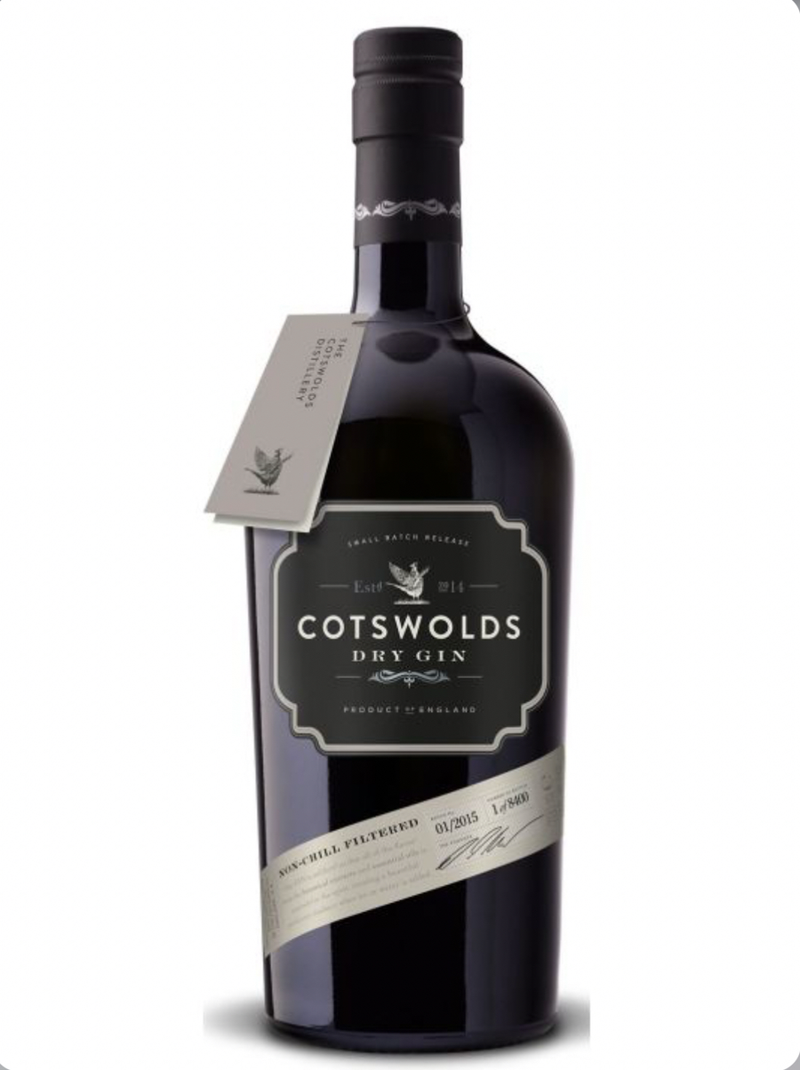 Dark purple-black glass bottle of Cotswolds Dry Gin (70cl), filled with clear gin. The bottle has a black cap and a matte black label with a cream framed panel in the center: "COTSWOLDS" in large white capitals at the top, "DRY GIN" below, a small white pheasant illustration, "Est. 2014" with a bird emblem, and "SMALL BATCH RELEASE" in smaller text. A diagonal silver foil band wraps around the bottle with "NON-CHILL FILTERED", "01/2015", batch number "1 of 8400", and a handwritten-style signature.