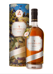 Tall cylindrical gift tube and amber whisky bottle of Cotswolds Harvest Series Amber Meadow Single Malt Whisky (70cl). The tube features a vibrant, impressionist-style wrap-around illustration of Cotswolds countryside in harvest season—golden fields, rolling hills, blue sky with clouds, wildflowers, and butterflies in warm blues, greens, yellows, and oranges. Text on the tube includes "THE HARVEST SERIES LIMITED RELEASE", "COTSWOLDS SINGLE MALT WHISKY", "AMBER MEADOW", and a decorative oak leaf emblem.