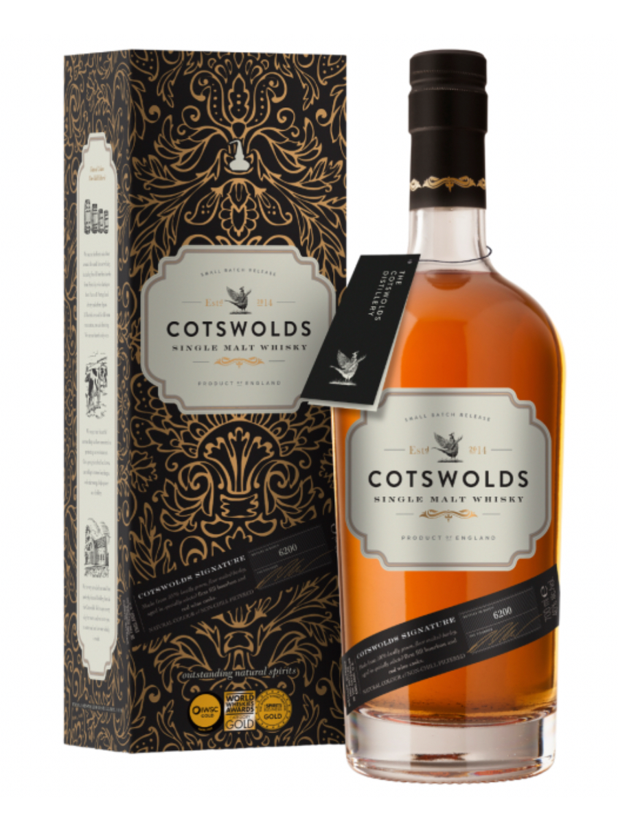 Dark navy gift tube and golden-amber whisky bottle of Cotswolds Signature Single Malt Whisky (70cl). The tall cylindrical tube is matte navy blue with intricate gold damask-style floral patterns and ornate text: "COTSWOLDS SINGLE MALT WHISKY", "SMALL BATCH RELEASE", "Est. 2014" with a pheasant emblem, and "PRODUCT OF ENGLAND". 