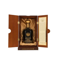 Luxurious wooden presentation box with open hinged doors revealing a bottle of Diplomático Ambassador Selección Rum (70cl) nestled on satin gold lining. The box is rich dark wood with brass hinges and "AMBASSADOR" engraved on the base. Inside, the bottle is deep black glass filled with rich dark amber rum, topped with an ornate gold cap and medallion featuring a bearded portrait (Don Juancho Nieto) in gold relief. The label is gold and black with elegant script reading "DIPLOMÁTICO AMBASSADOR Selección"