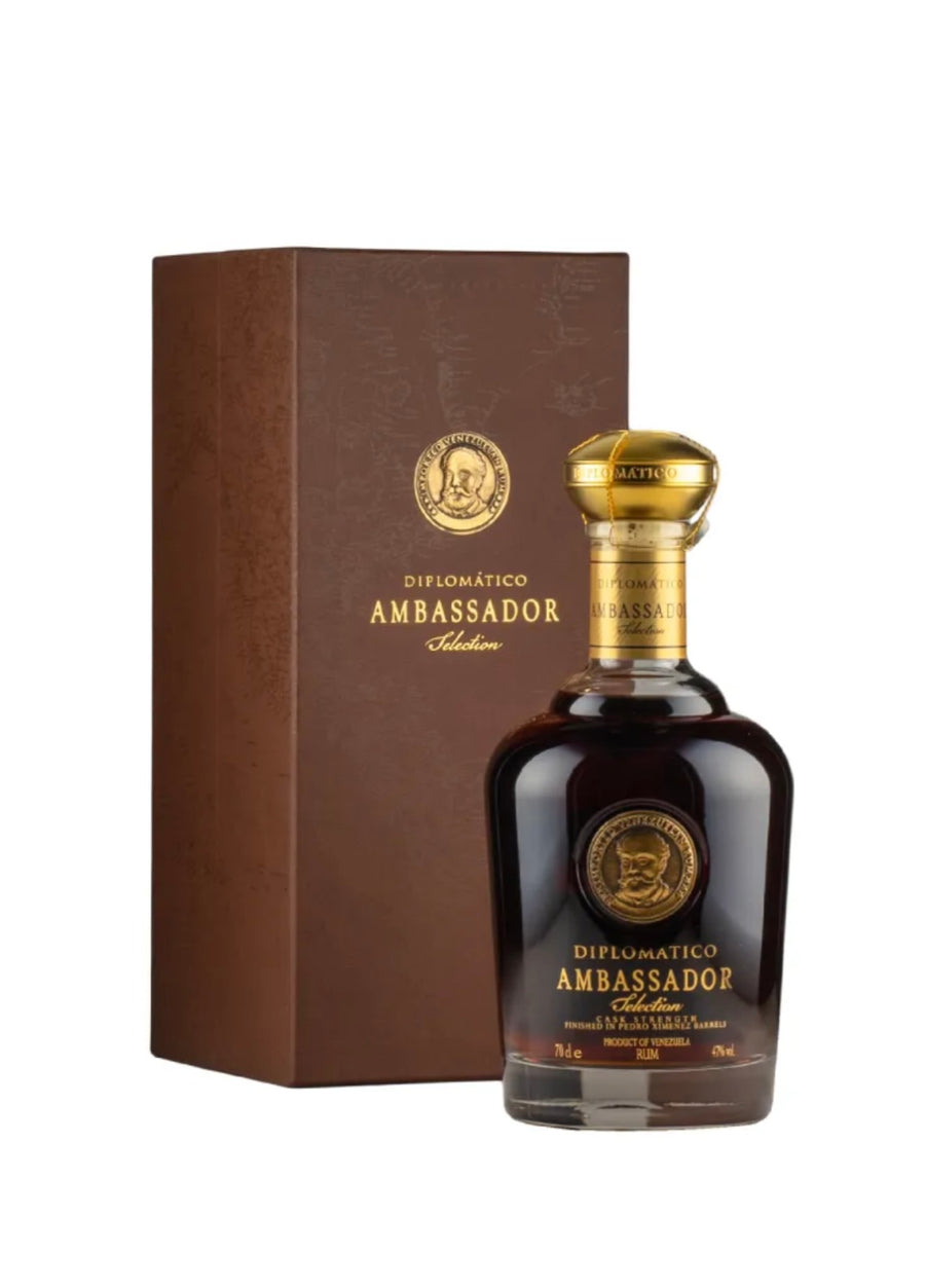 Dark brown gift box and deep amber rum bottle of Diplomático Ambassador Selección Rum (70cl / 700ml). The tall rectangular box is matte brown with subtle heritage patterns and gold-embossed text reading "DIPLOMÁTICO AMBASSADOR" and "Selección". The bottle has a luxurious dark glass body filled with rich, deep amber liquid, a gold ornate cap with the Diplomático emblem, and a prominent circular gold medallion label featuring a portrait of a bearded man (Don Juancho Nieto) in sepia tones