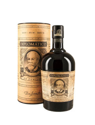 Tall cylindrical cardboard tube gift box and dark bottle of Diplomático Selección de Familia Rum (70cl). The box is kraft-brown with vintage barrel-like woodgrain texture, printed in black and gold with "RON - RUM - RHUM" repeated at the top, "DIPLOMÁTICO" prominently centered, "SELECCIÓN DE FAMILIA" below, and "Don Juancho" signature at the base. The bottle is deep dark green glass with rich amber rum inside, featuring a black cap, a cream-and-black vintage-style label with ornate gold borders