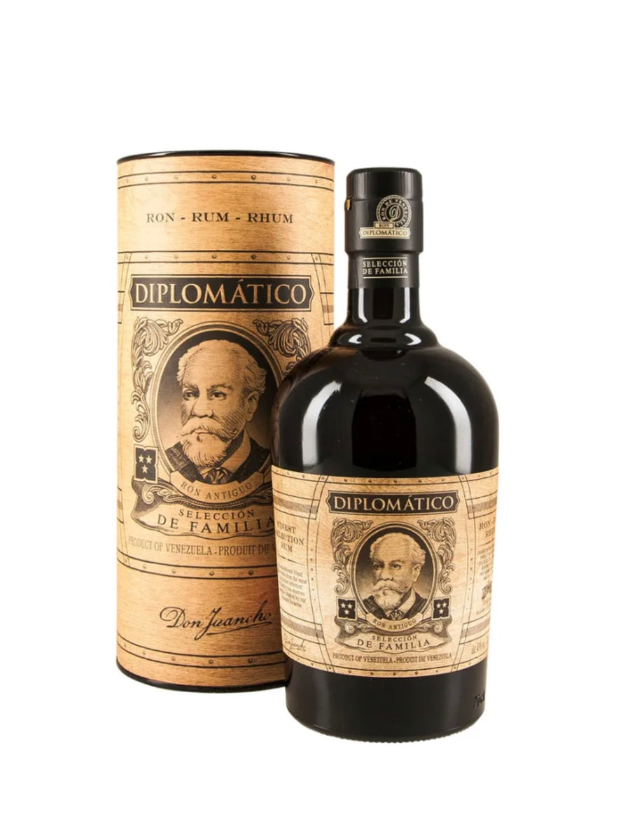 Tall cylindrical cardboard tube gift box and dark bottle of Diplomático Selección de Familia Rum (70cl). The box is kraft-brown with vintage barrel-like woodgrain texture, printed in black and gold with "RON - RUM - RHUM" repeated at the top, "DIPLOMÁTICO" prominently centered, "SELECCIÓN DE FAMILIA" below, and "Don Juancho" signature at the base. The bottle is deep dark green glass with rich amber rum inside, featuring a black cap, a cream-and-black vintage-style label with ornate gold borders