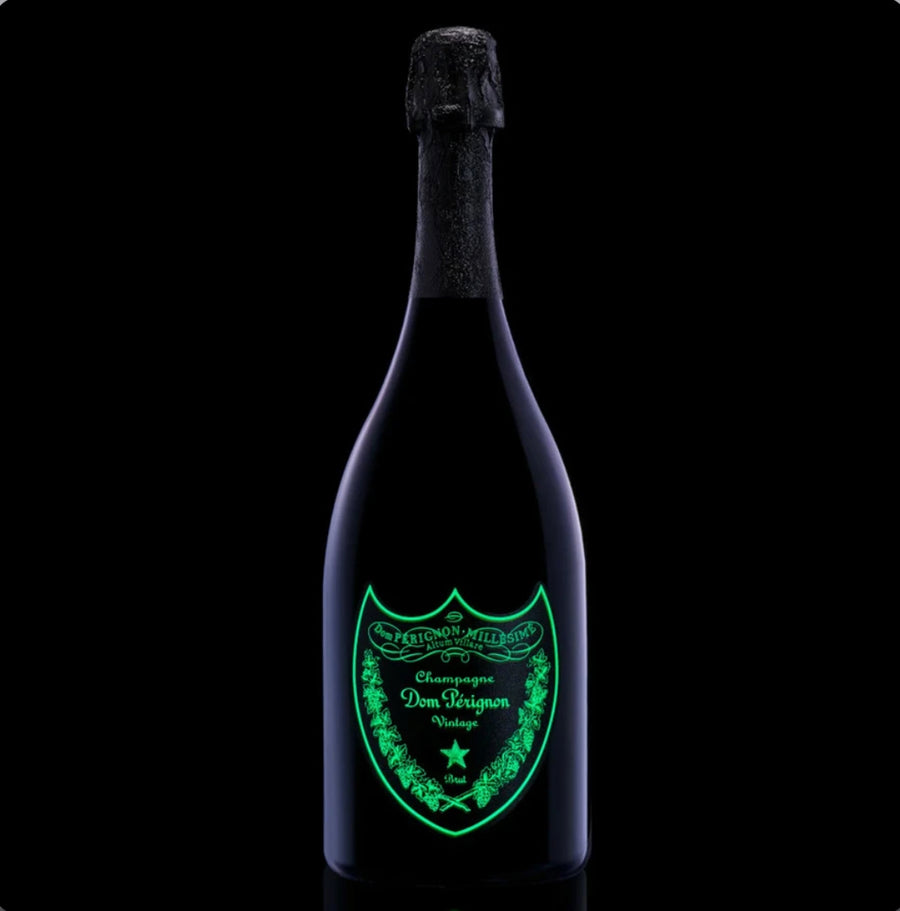 Dom Pérignon Luminous Label Vintage 2013 Champagne 75cl bottle glowing in the dark, with a neon green illuminated shield-shaped label featuring ornate vine details, “Champagne Dom Pérignon Vintage” text, a star emblem, and “Brut” designation on a sleek black glass silhouette against a dark background.