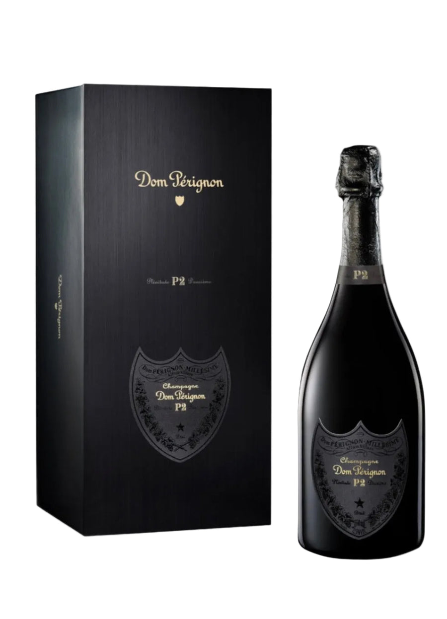 A bottle of Dom Perignon P2 Champagne next to its black presentation box, both featuring gold lettering and the classic shield logo, set against a clean white background with a subtle reflection.