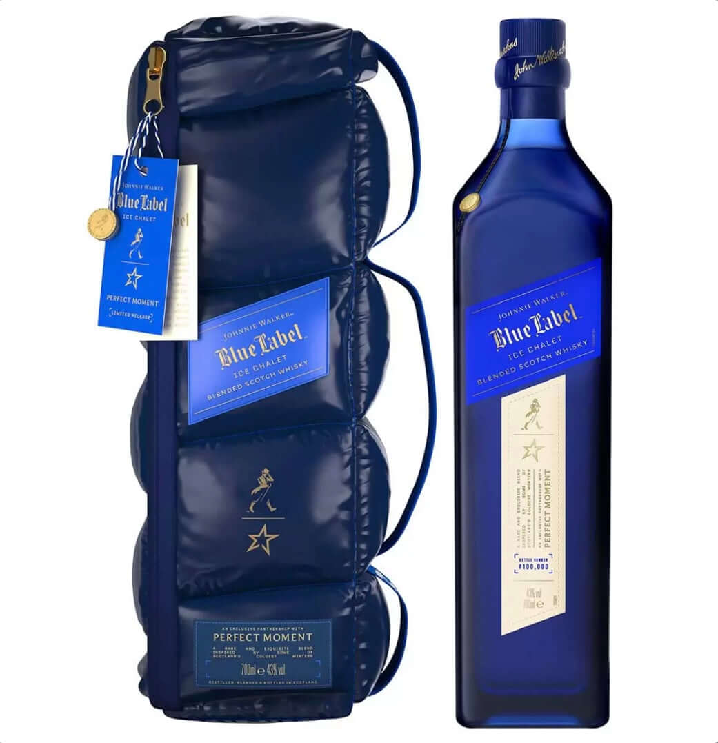 Navy blue puffer-style insulated bottle bag encasing a bottle of Johnnie Walker Blue Label Ice Chalet limited-edition blended Scotch whisky, designed in collaboration with Perfect Moment. The reversible, quilted bag has a glossy, padded, down-like texture with horizontal stitching, a detachable crossbody strap, gold zipper pull, and features the iconic gold striding Johnnie Walker figure and a gold North Star emblem repeated along the front. 