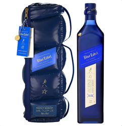 Navy blue puffer-style insulated bottle bag encasing a bottle of Johnnie Walker Blue Label Ice Chalet limited-edition blended Scotch whisky, designed in collaboration with Perfect Moment. The reversible, quilted bag has a glossy, padded, down-like texture with horizontal stitching, a detachable crossbody strap, gold zipper pull, and features the iconic gold striding Johnnie Walker figure and a gold North Star emblem repeated along the front. 