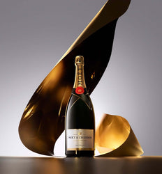 A bottle of Moët & Chandon Non Volontaire sparkling wine stands on a dark surface against a gray gradient background, framed by sculptural flowing gold metallic drapes.