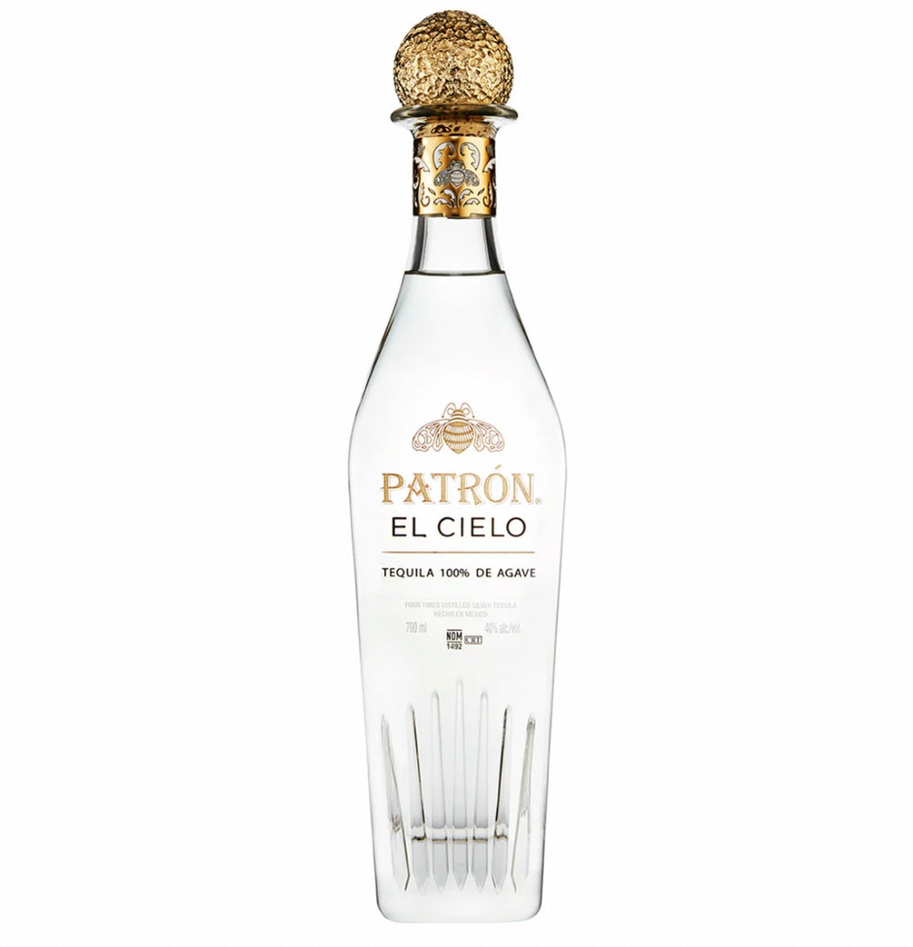 Clear glass bottle of Patrón El Cielo four-times distilled luxury blanco tequila with a distinctive tall, elegant shape featuring vertical etched lines at the base resembling agave leaves or crystal facets. The bottle contains crystal-clear liquid and has a prominent gold foil capsule with intricate embossed bee and floral motifs.