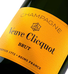 Close-up of a Veuve Clicquot Brut champagne bottle label, showing the brand name, 'Champagne', and 'BRUT' on a bright yellow-orange label against a dark bottle background.