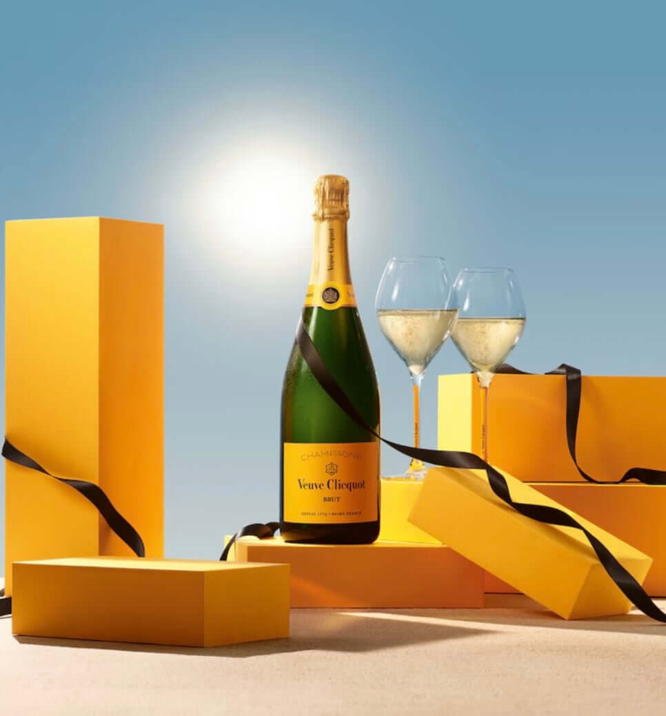 A bottle of Veuve Clicquot champagne stands between two filled wine glasses, surrounded by bright yellow gift boxes and black ribbons against a blue sky background with a bright sun glow.