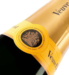 Close-up of a champagne bottle with a gold foil label and a round black-and-gold crest seal. The brand name 'Veuve' is partially visible on the gold wrap against a white background.