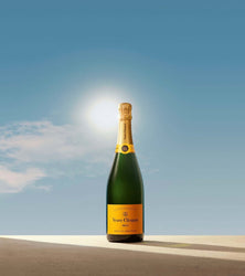 A bottle of Veuve Clicquot champagne stands upright on a sunlit surface against a bright blue sky with the sun shining behind it.