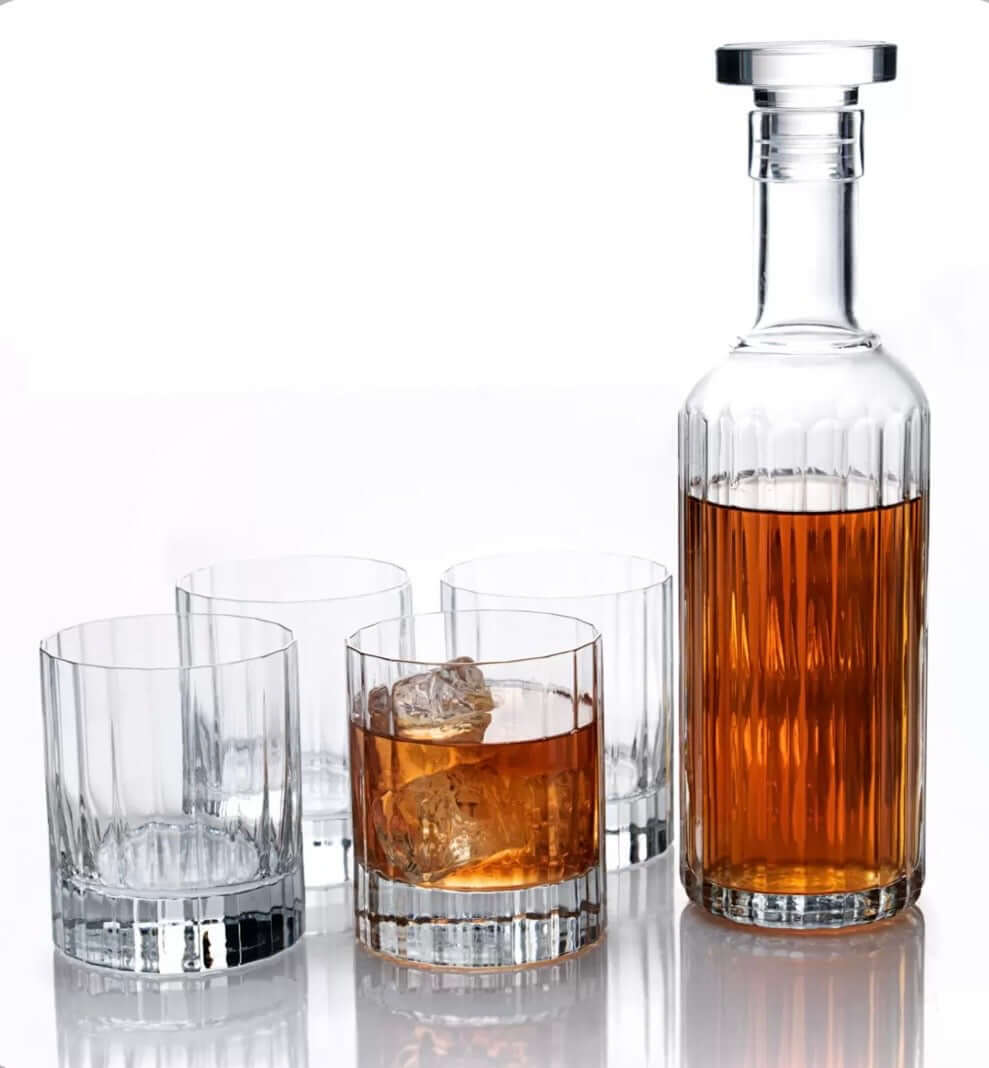 A clear glass decanter with a stopper filled with amber liquor, shown beside four matching clear lowball glasses on a white reflective surface.