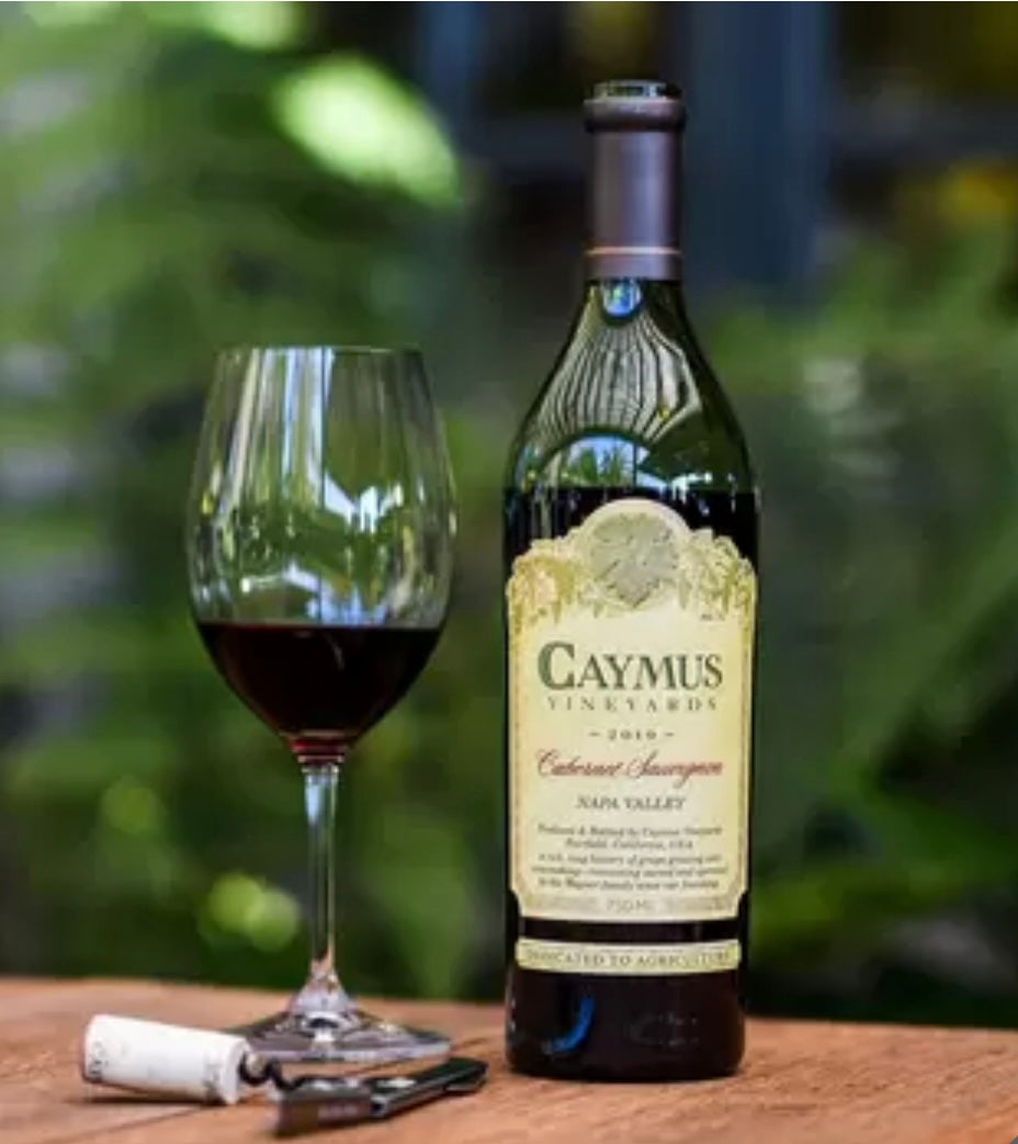 Caymus Vineyards Cabernet Sauvignon 2022 75cl 50th Anniversary bottle on a garden table with a wine opener, set in an outdoor yard.
